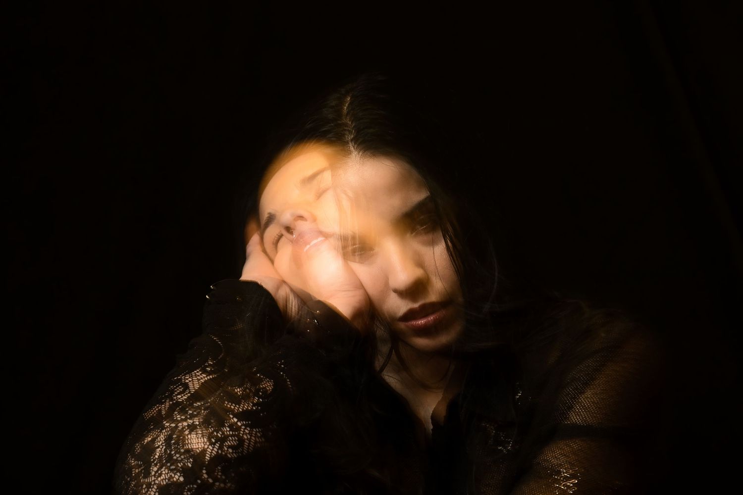 Blurred long exposure portrait of a woman with hands on face in dark setting.