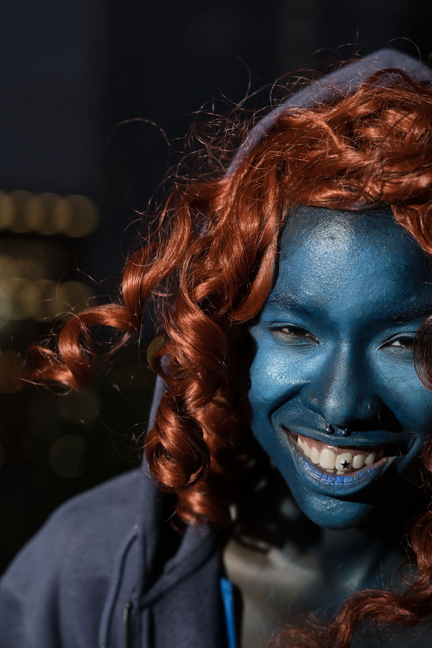 Smiling person wearing blue face paint and red curly wig with nose ring outdoors.