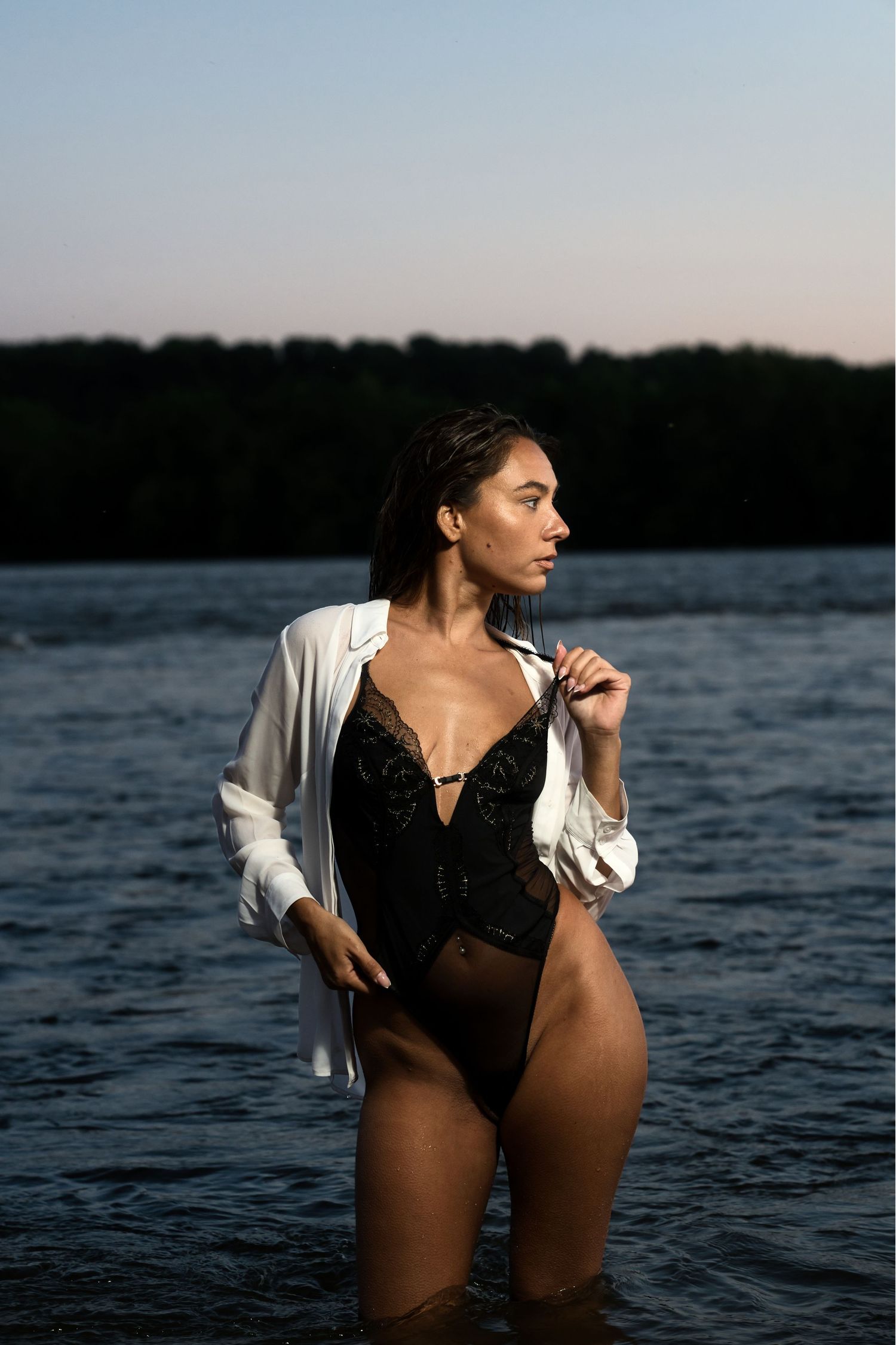 Woman standing in a lake wearing a black lace bodysuit and open white shirt at dusk.