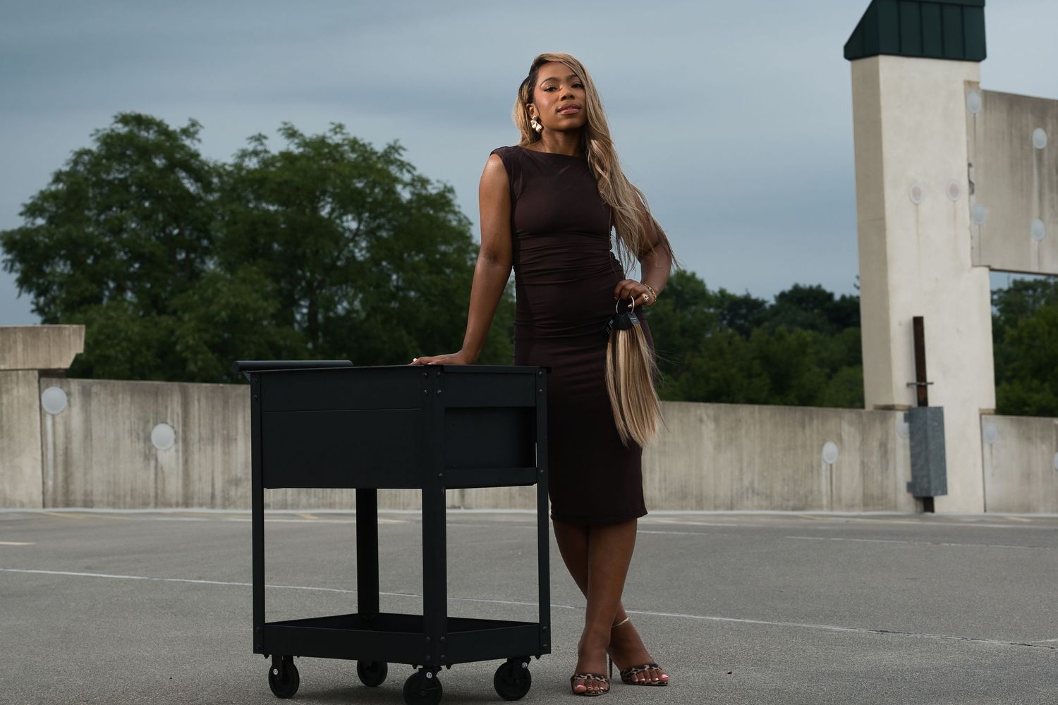 Woman in a brown dress standing beside a black rolling cart on a rooftop.
