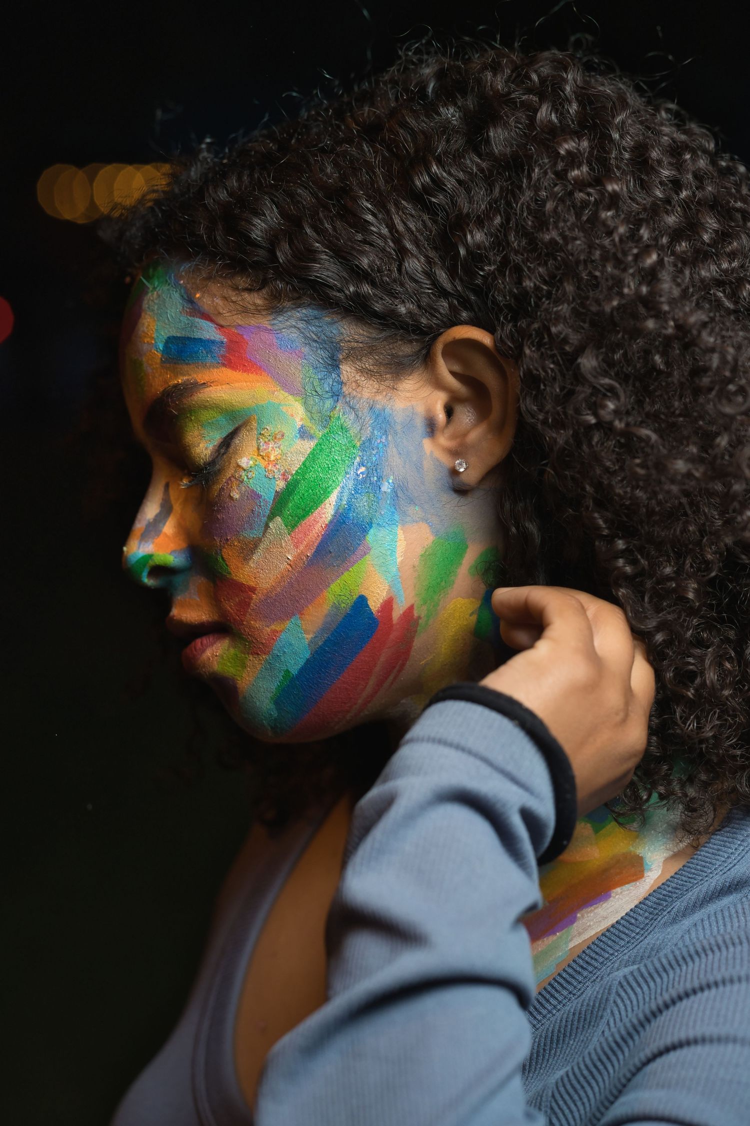 Young woman with curly hair and colorful face paint posing with eyes closed in dark setting.