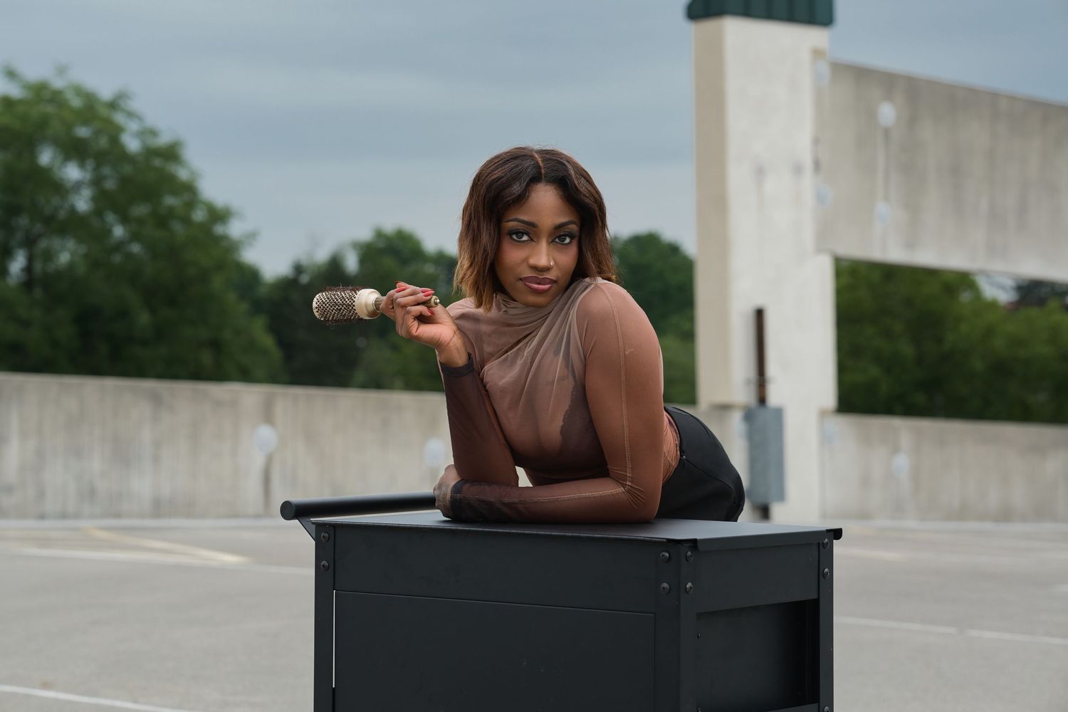 Woman leaning on a black cart holding a microphone on a rooftop parking lot.