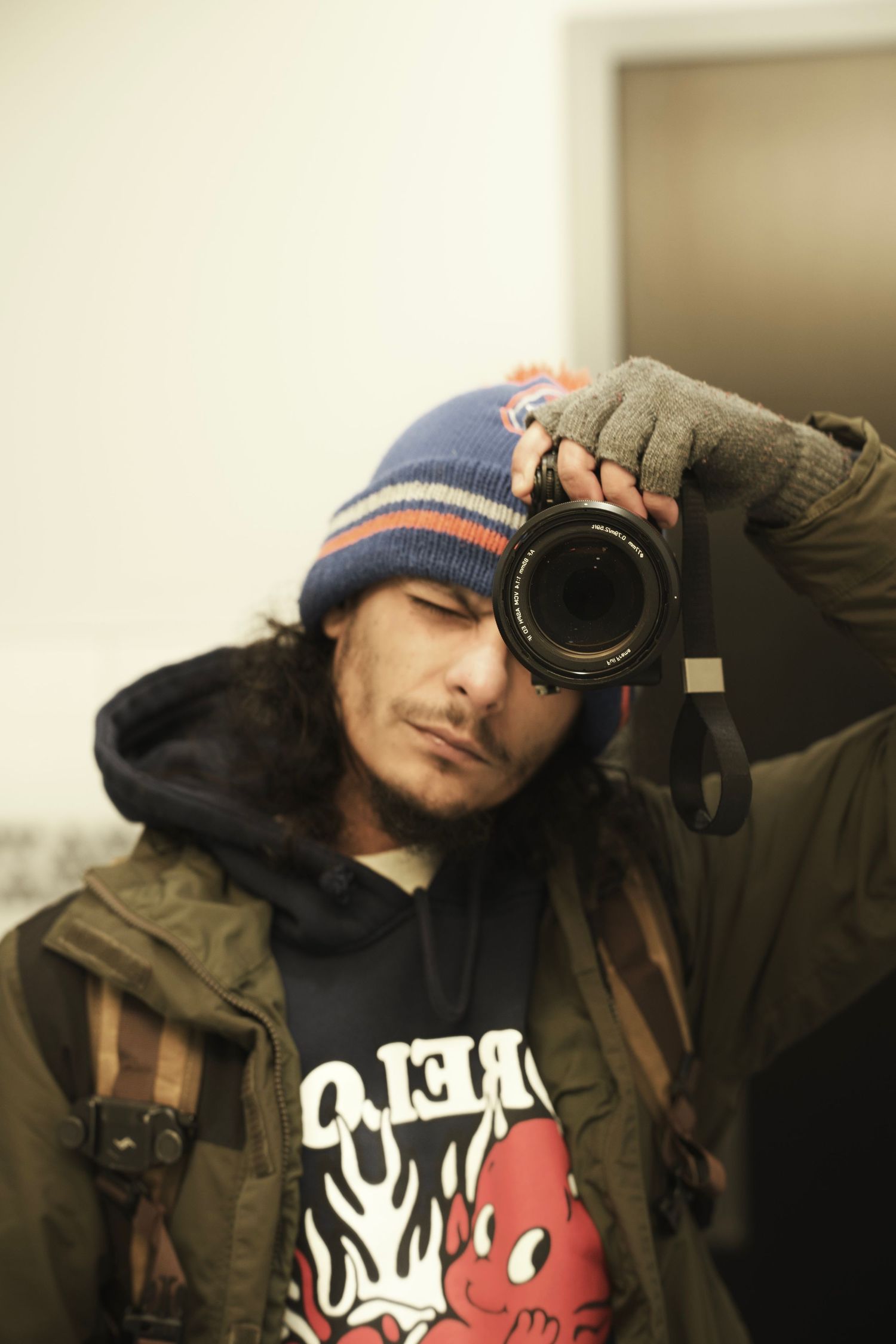 Photographer in a beanie and layered jacket holds a DSLR camera up to his face indoors.