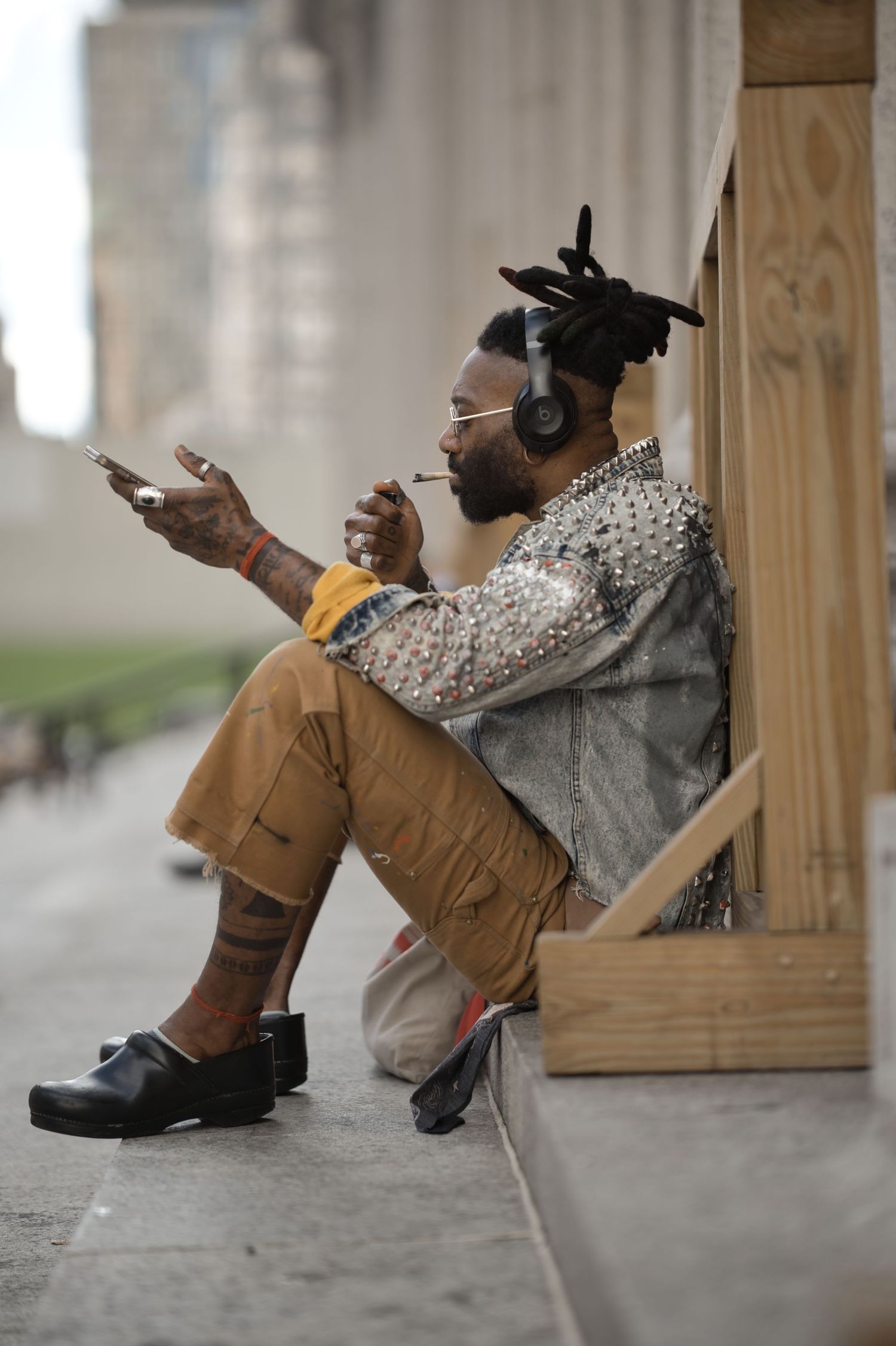 Man with dreadlocks wearing studded jacket, headphones, sitting on ledge using phone while smoking in urban setting.