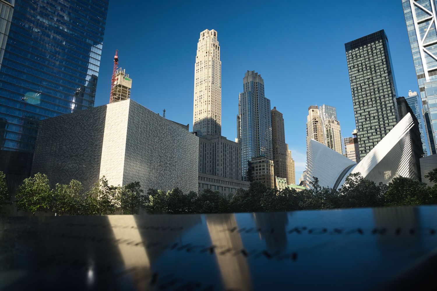 9/11 Memorial reflecting pool with NYC skyline and One World Trade Center in background on a clear sunny day.