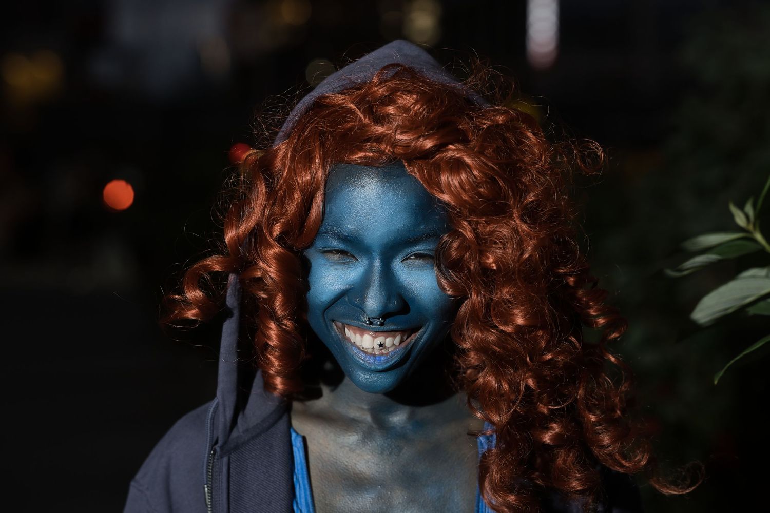 Person in blue face paint and red curly wig grinning outdoors at night.
