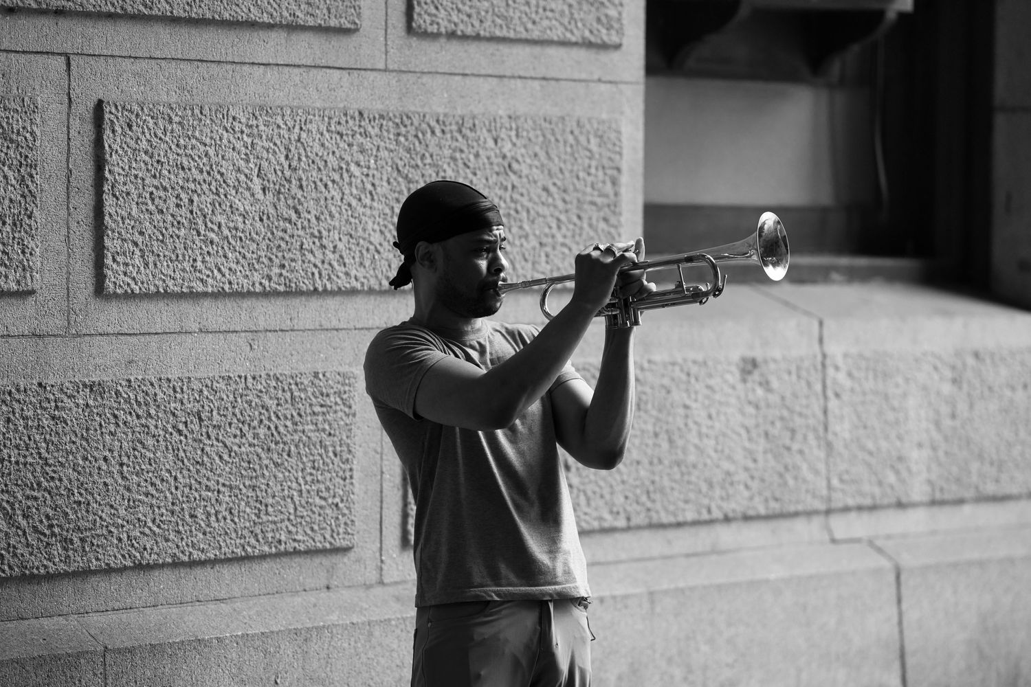 Black and white photo of a man playing trumpet against a stone wall outdoors.