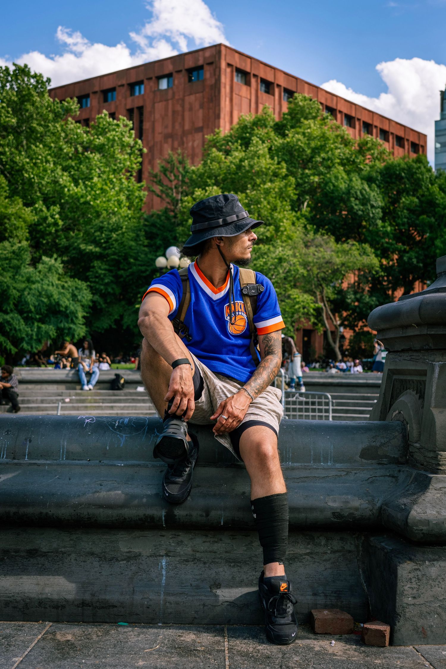 Tattooed man in blue Knicks jersey and bucket hat sitting on fountain ledge outdoors.