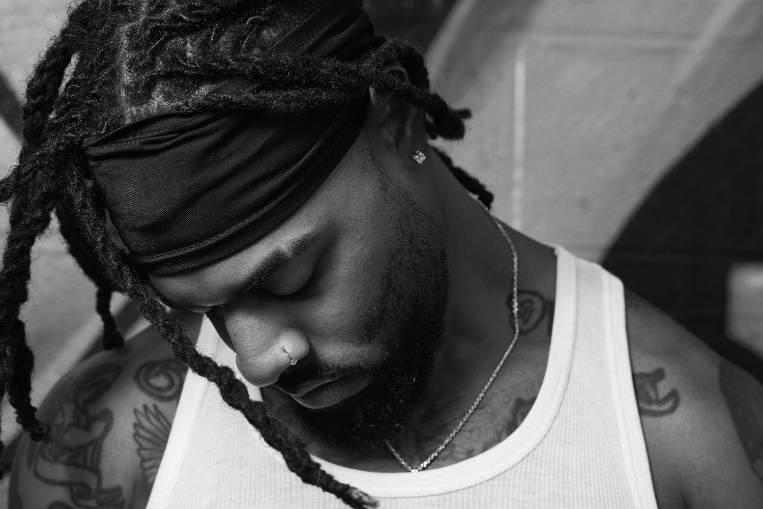 Black and white portrait of a man with dreadlocks and a headband wearing a white tank top with tattoos.