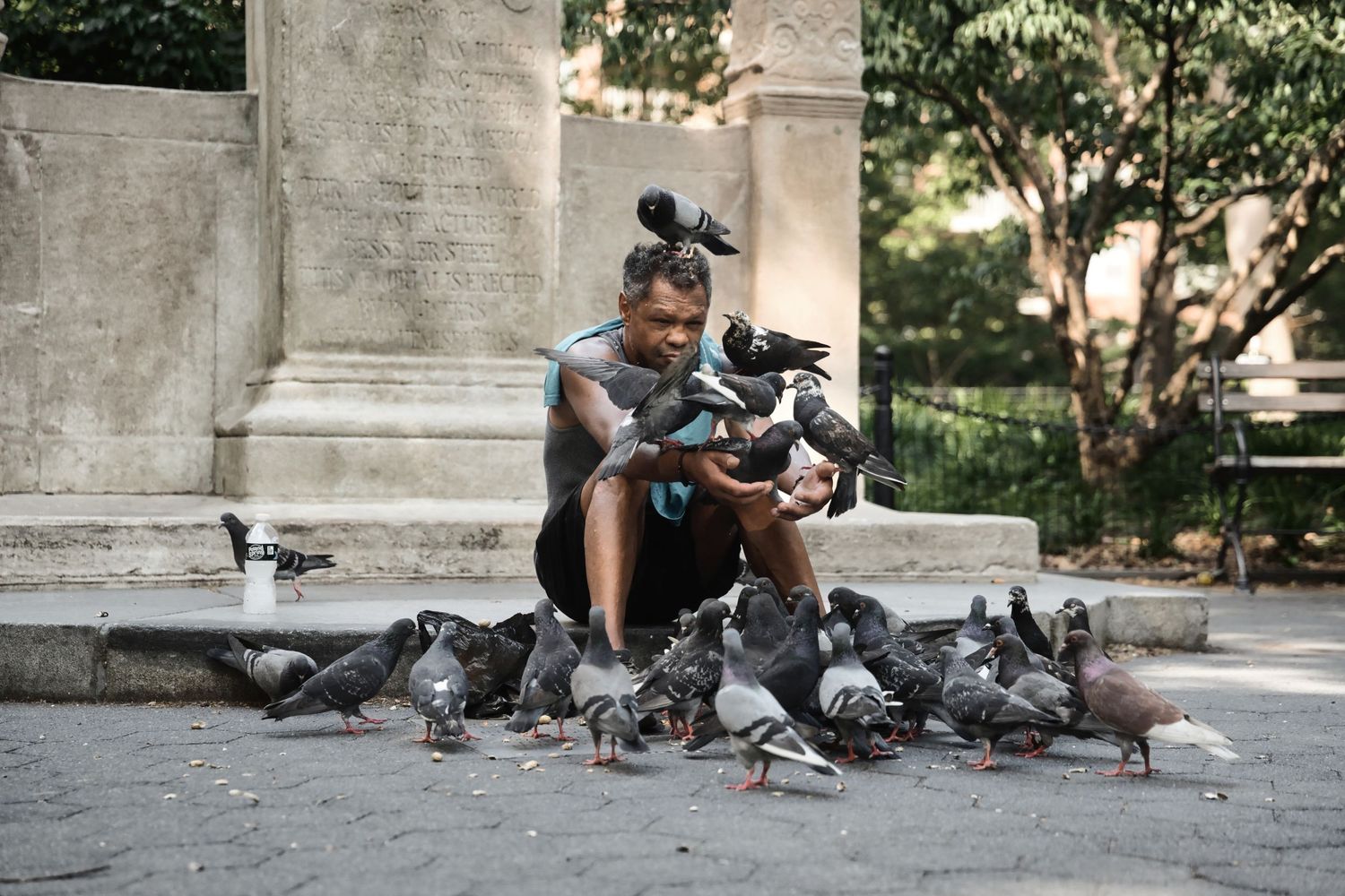 A man sits outdoors feeding a large flock of pigeons perched on his arms and gathered around his feet in a park.