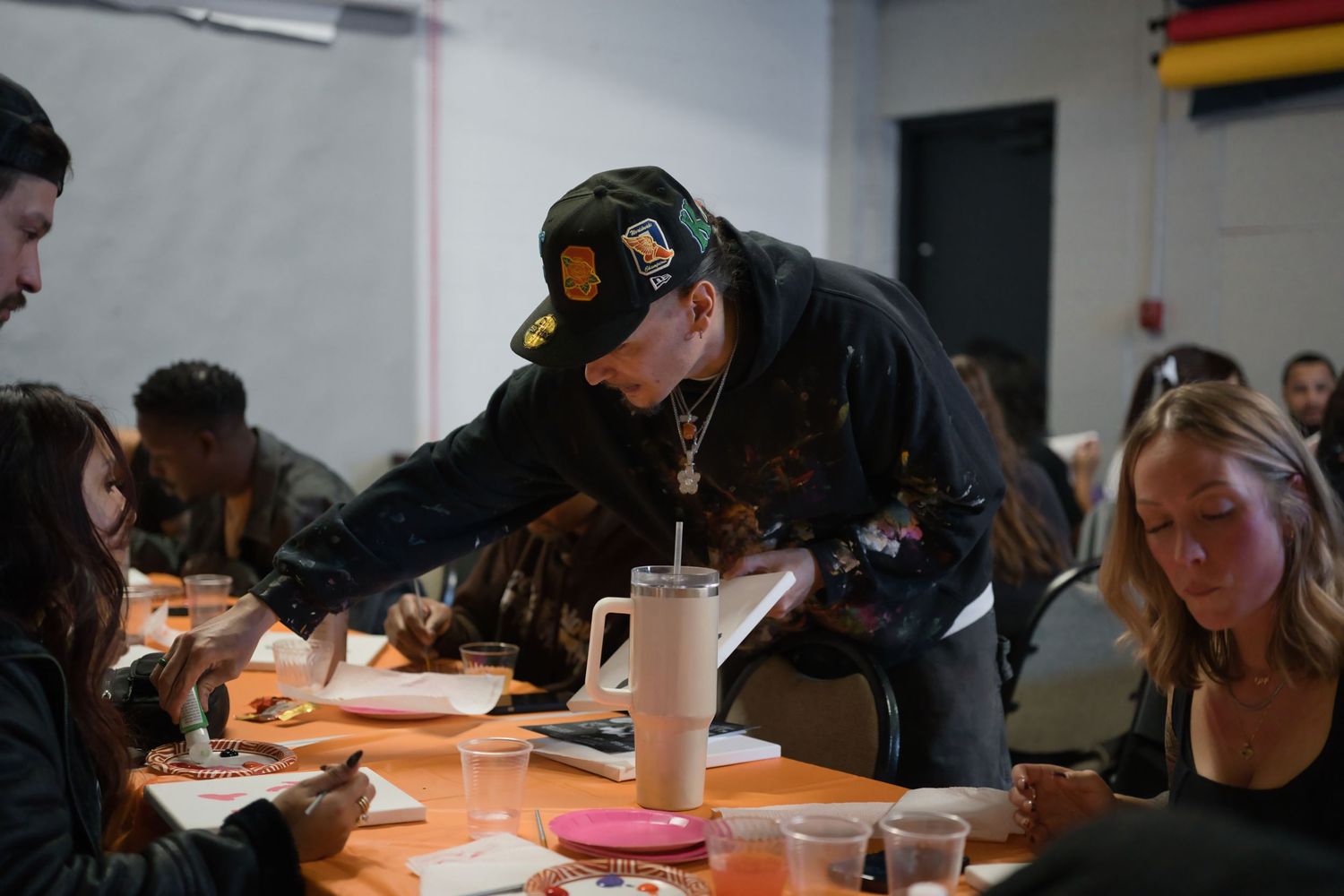 Person in military cap leans over long table helping seated participants at indoor group activity.