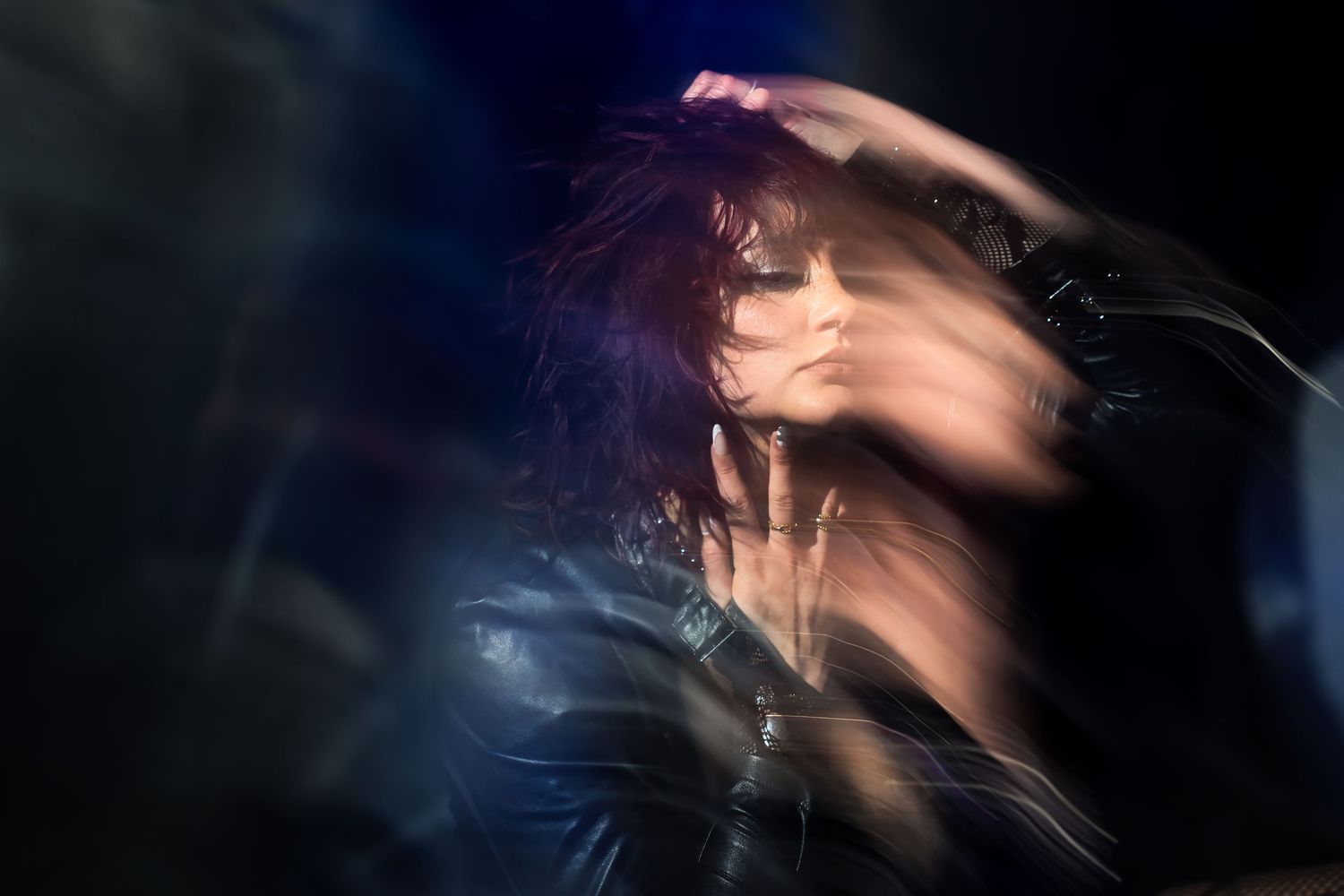A woman in a leather jacket headbanging with motion blur effect in dramatic blue stage lighting.