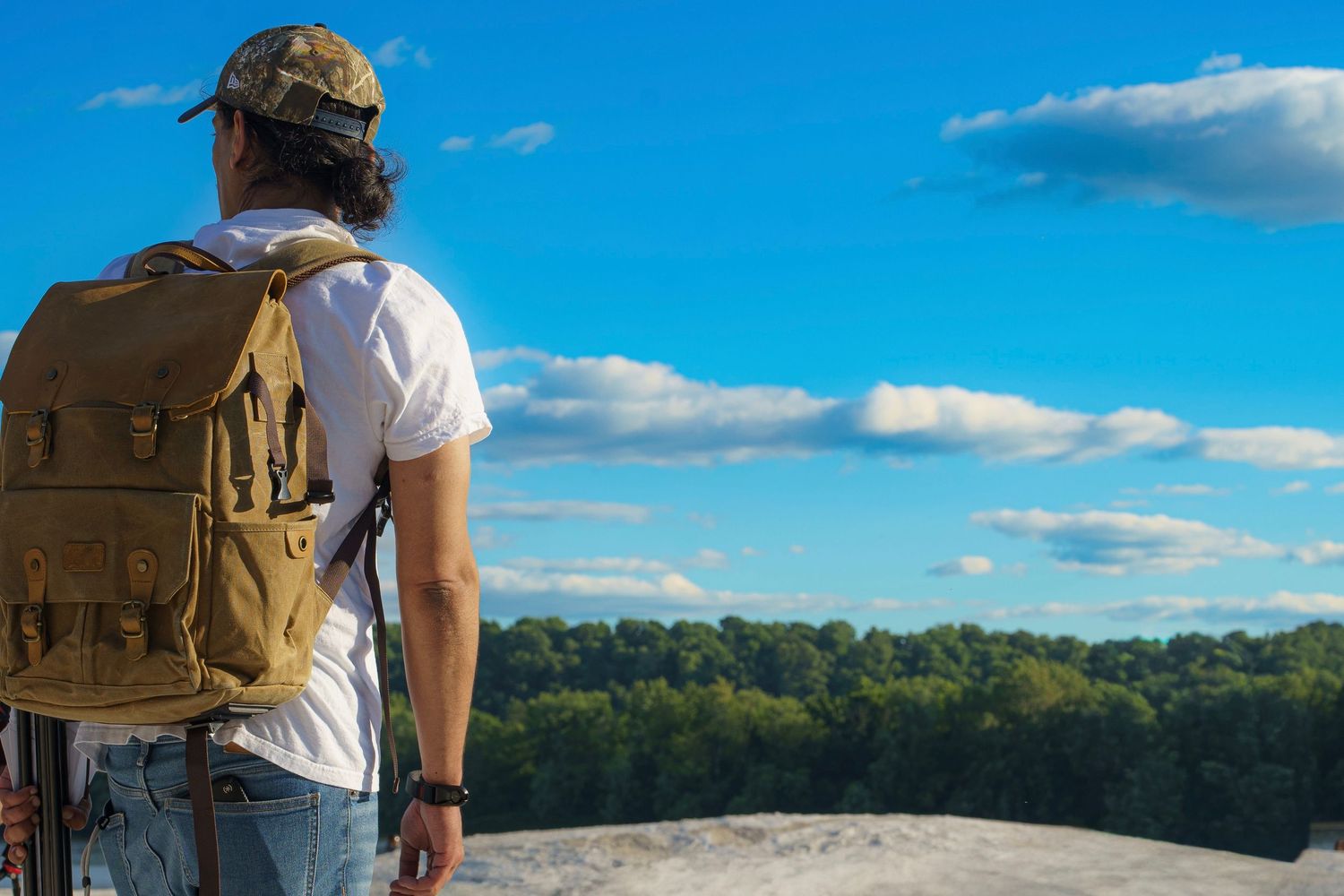 Hiker with tan backpack standing on rocky overlook admiring scenic forest and blue sky view.