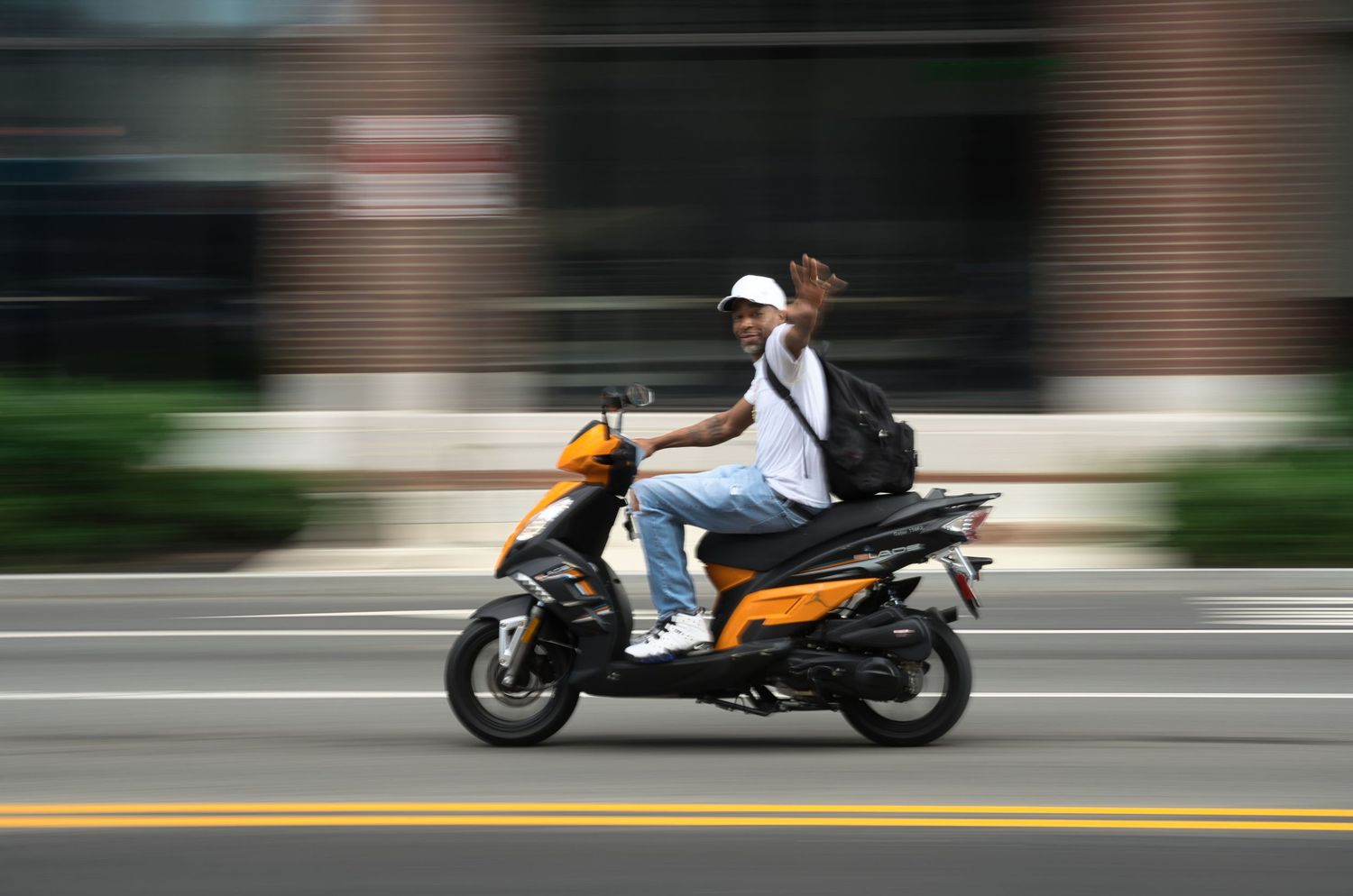 Person riding an orange and black motor scooter on a city street with motion blur background.