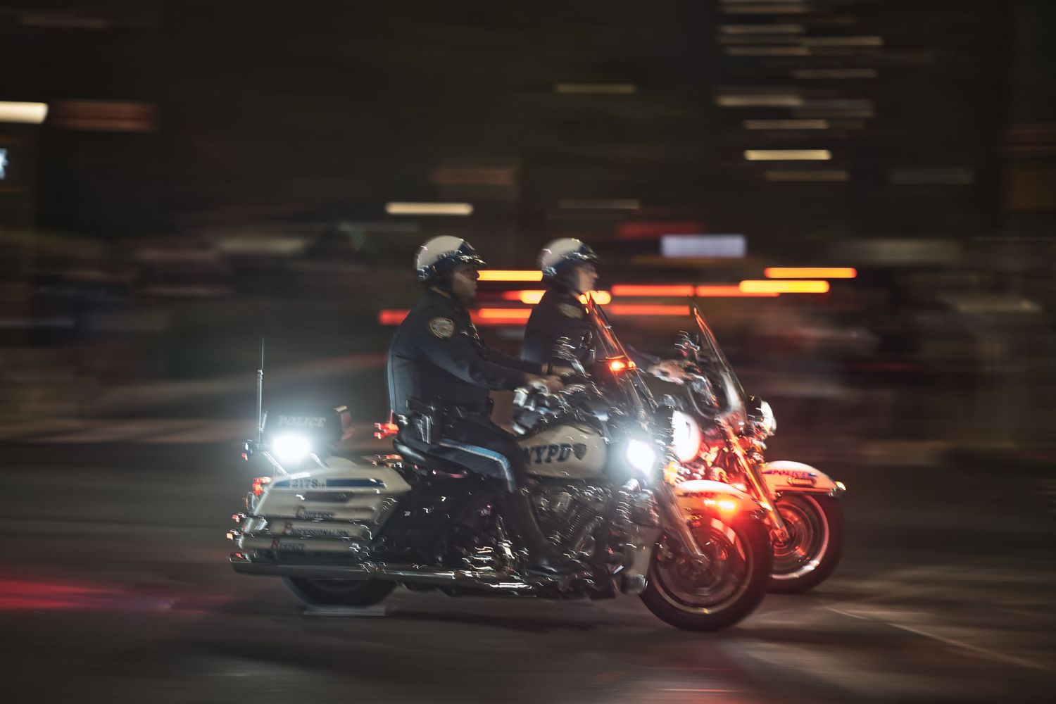 Two motorcyclists ride through a city street at night, with motion blur and bright lights creating a dynamic urban scene.
