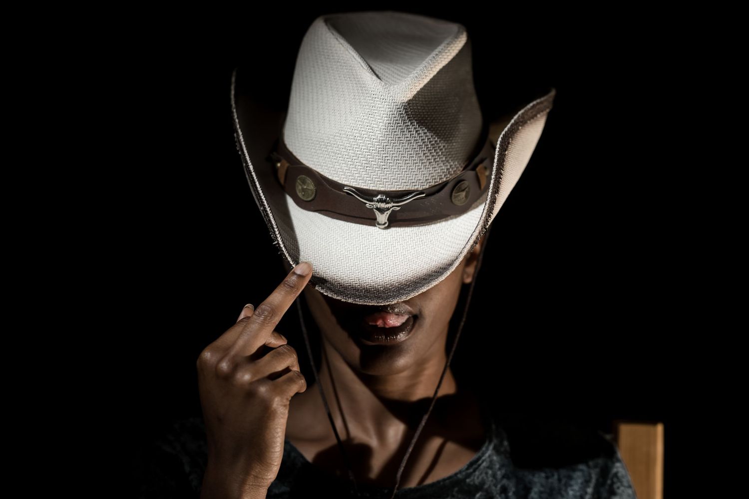 Person wearing a white cowboy hat tilted forward, pointing finger up against a dark background.