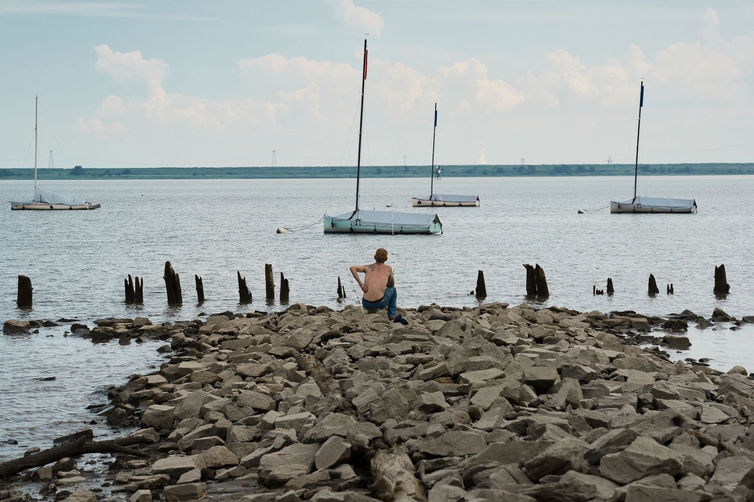 Person sitting on rocky shoreline watching sailboats on calm water under cloudy sky.