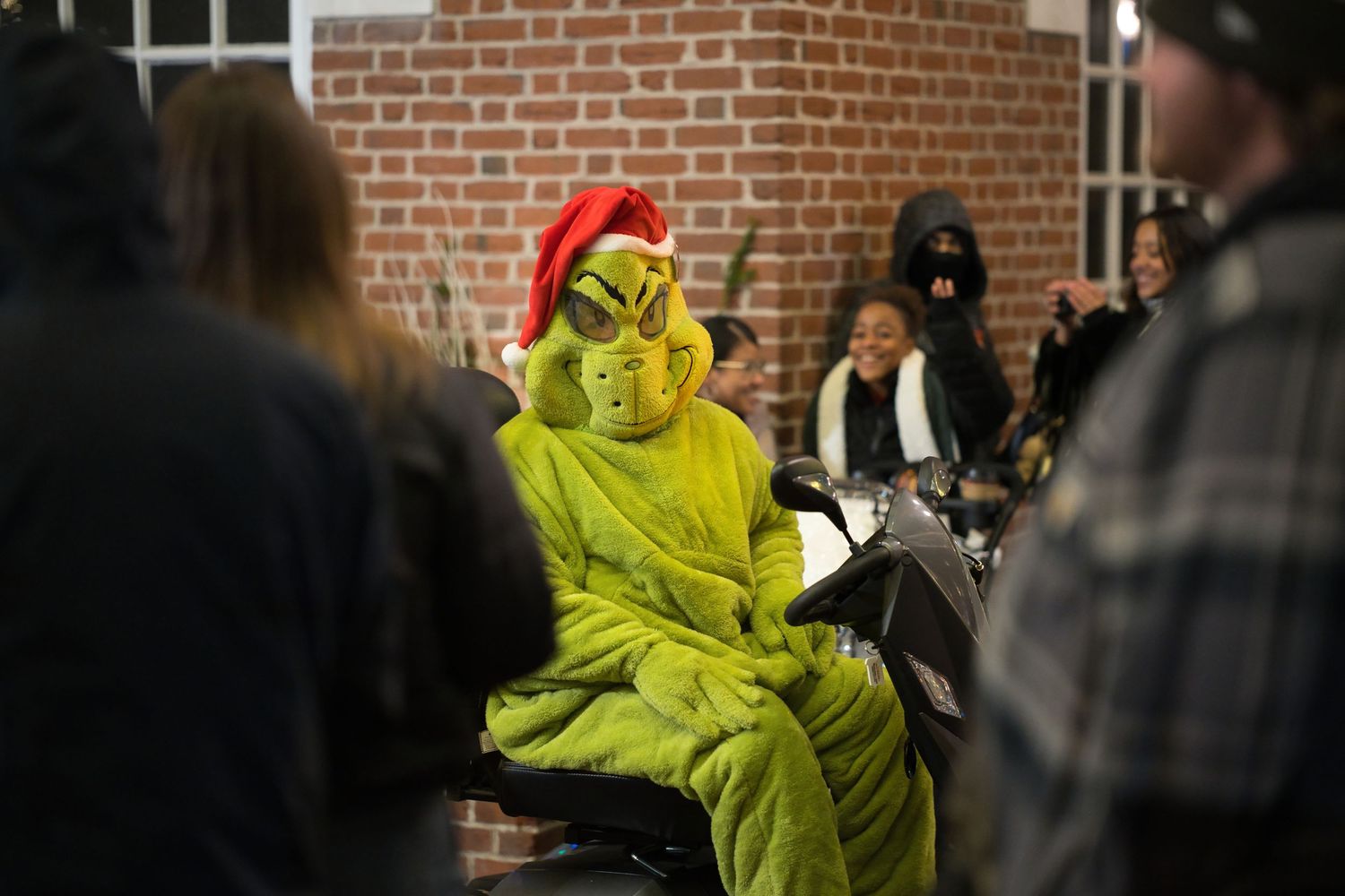Person dressed in Grinch costume with Santa hat sits on motorcycle at Christmas event.