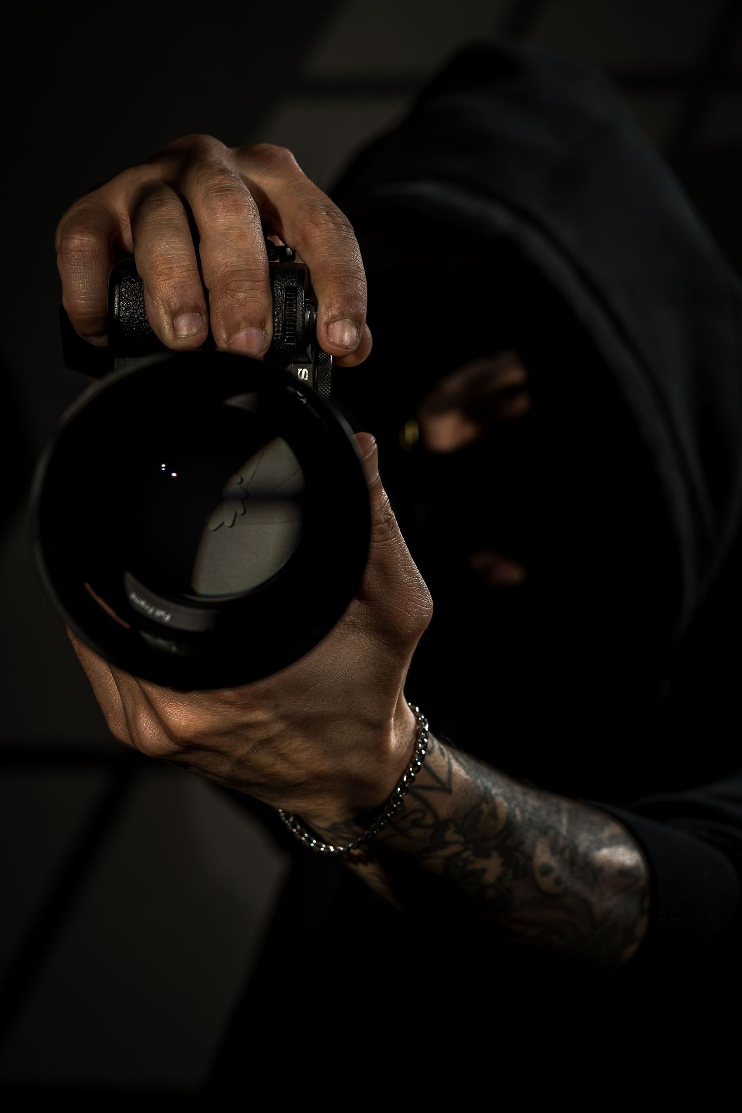 Tattooed photographer in black hoodie and mask holding a professional camera lens in dramatic lighting.