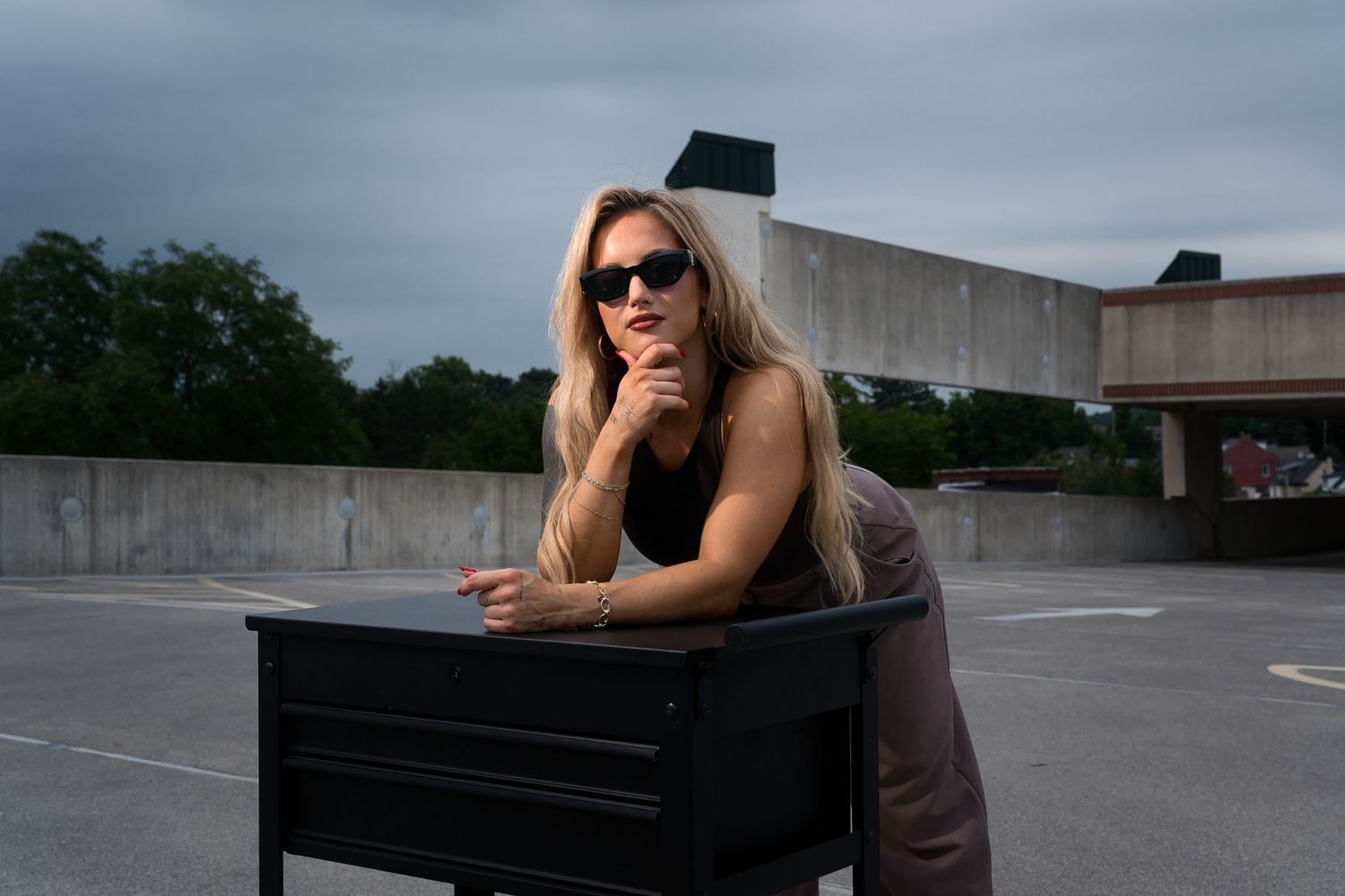 Blonde woman in sunglasses leaning on a black desk on a cloudy rooftop.
