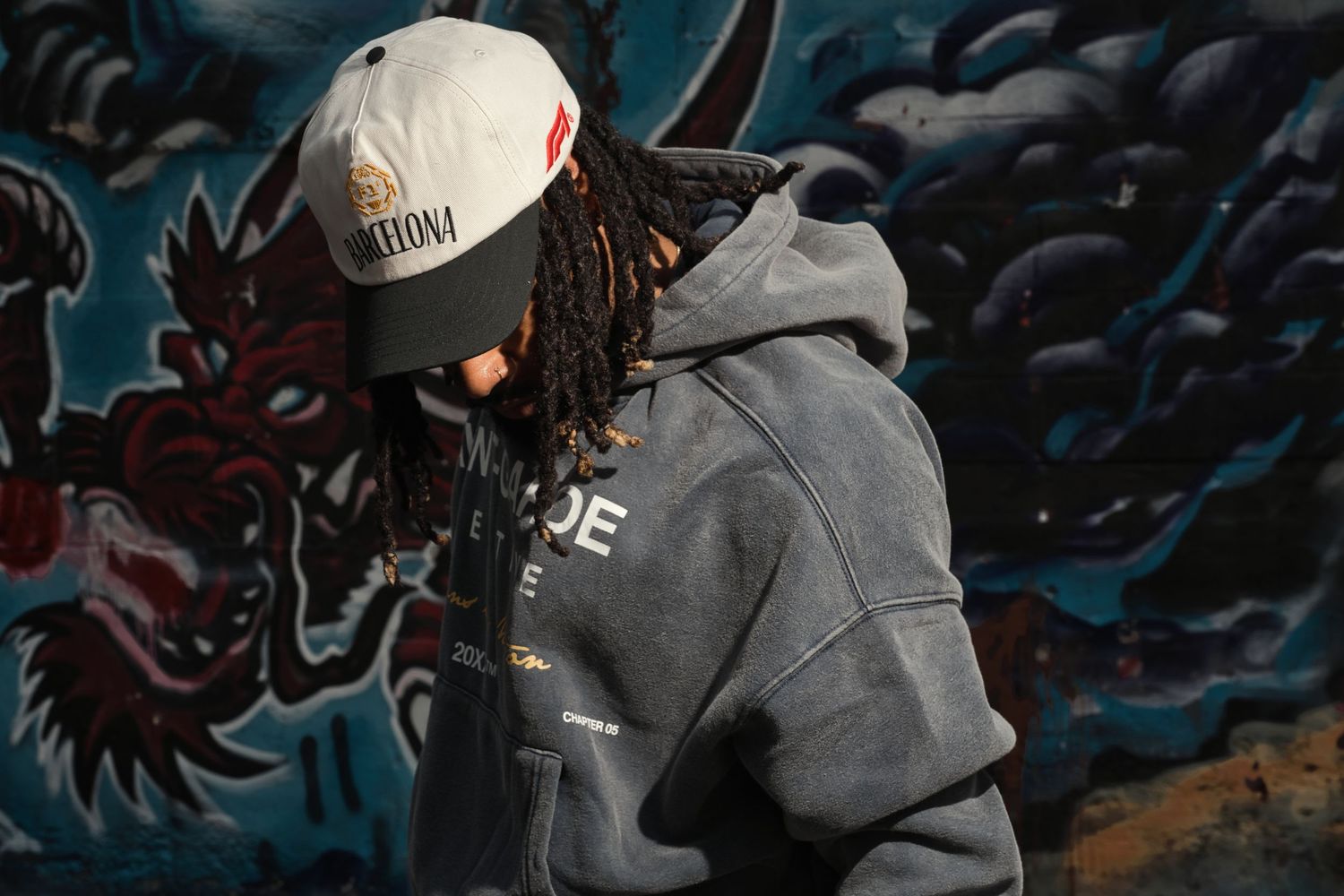 Man with dreadlocks wearing a gray hoodie and cap posing in front of a colorful dragon mural.