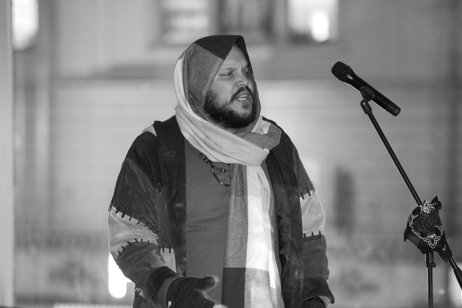 Black and white portrait of bearded man in headscarf speaking at microphone.