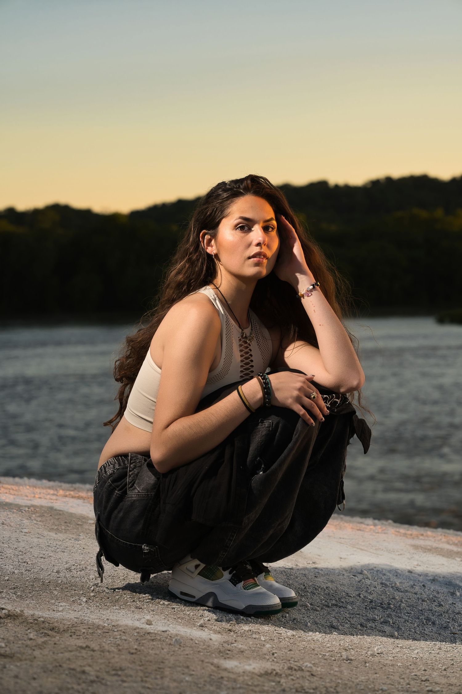 Young woman with long dark hair crouching outdoors near a lake at sunset in black pants and white tank top.