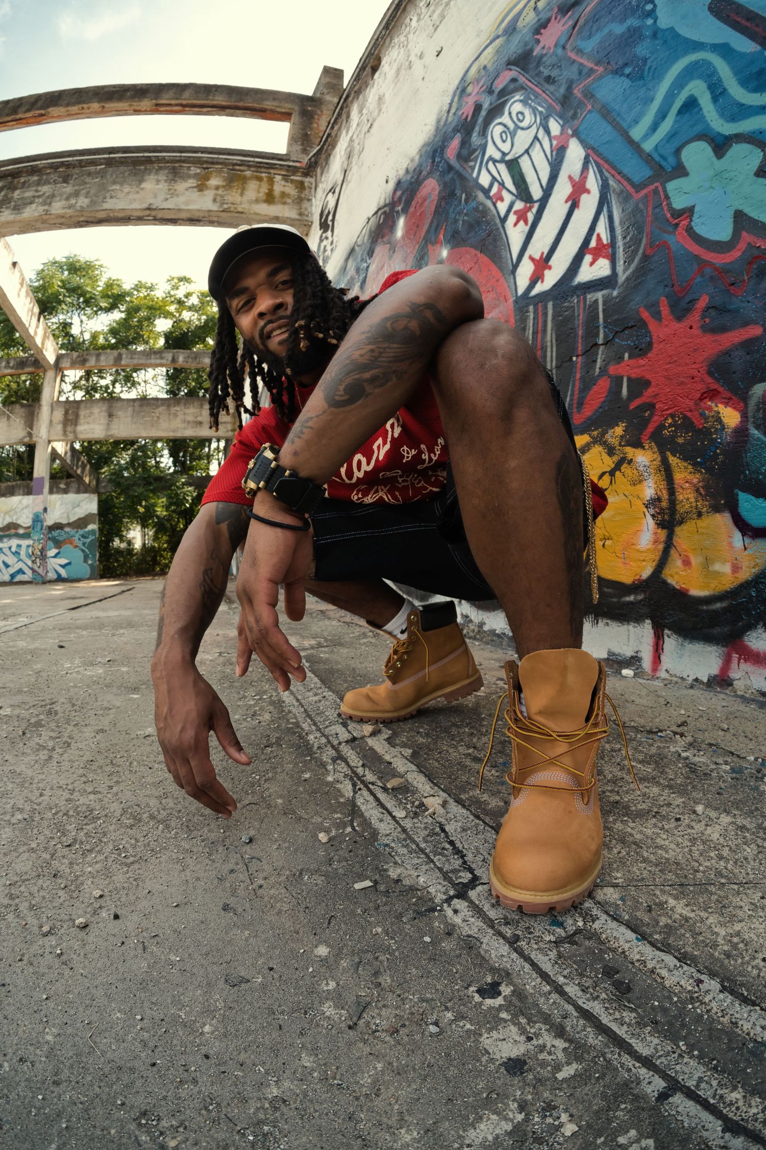 Man with dreadlocks crouching in tan boots and red shirt in front of vibrant graffiti wall.
