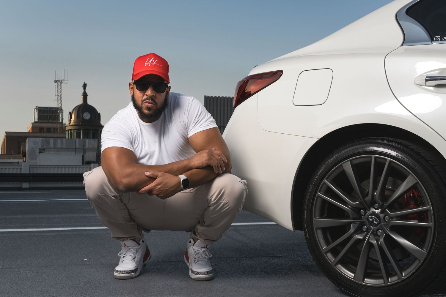 Man in red cap squatting beside white luxury car on rooftop parking lot.