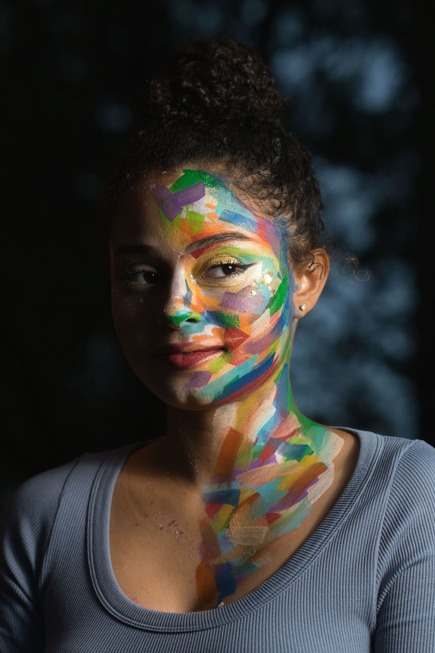 Portrait of a young woman with multicolored face paint smiling softly against a dark background.