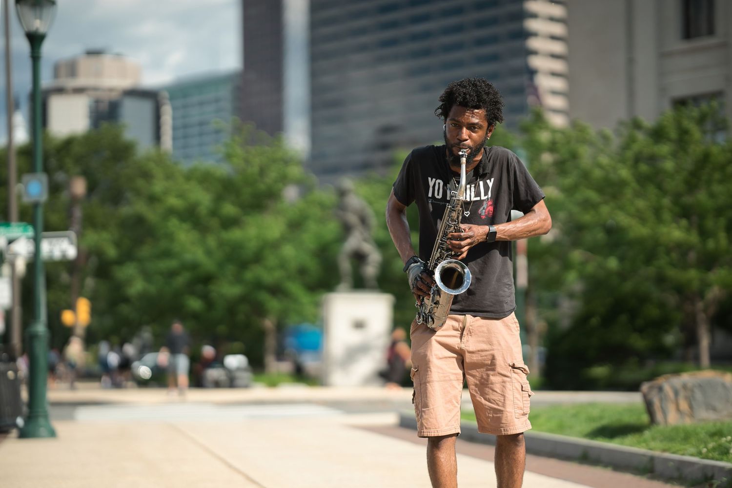 Young man playing saxophone on a city sidewalk wearing a black t-shirt and khaki shorts with buildings in background.