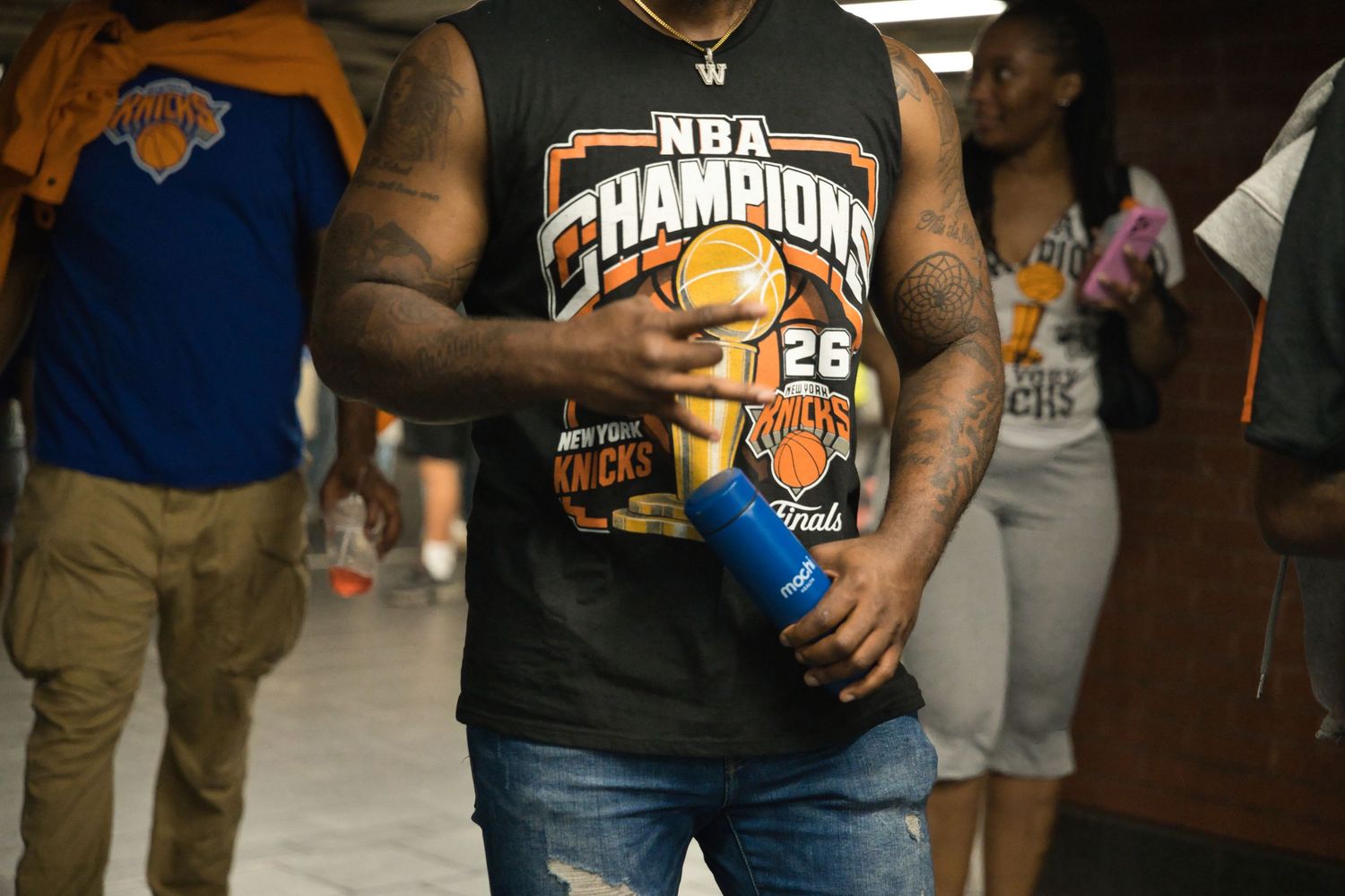 Man wearing NBA Champions New York Knicks shirt holding blue bottle in crowded indoor venue.