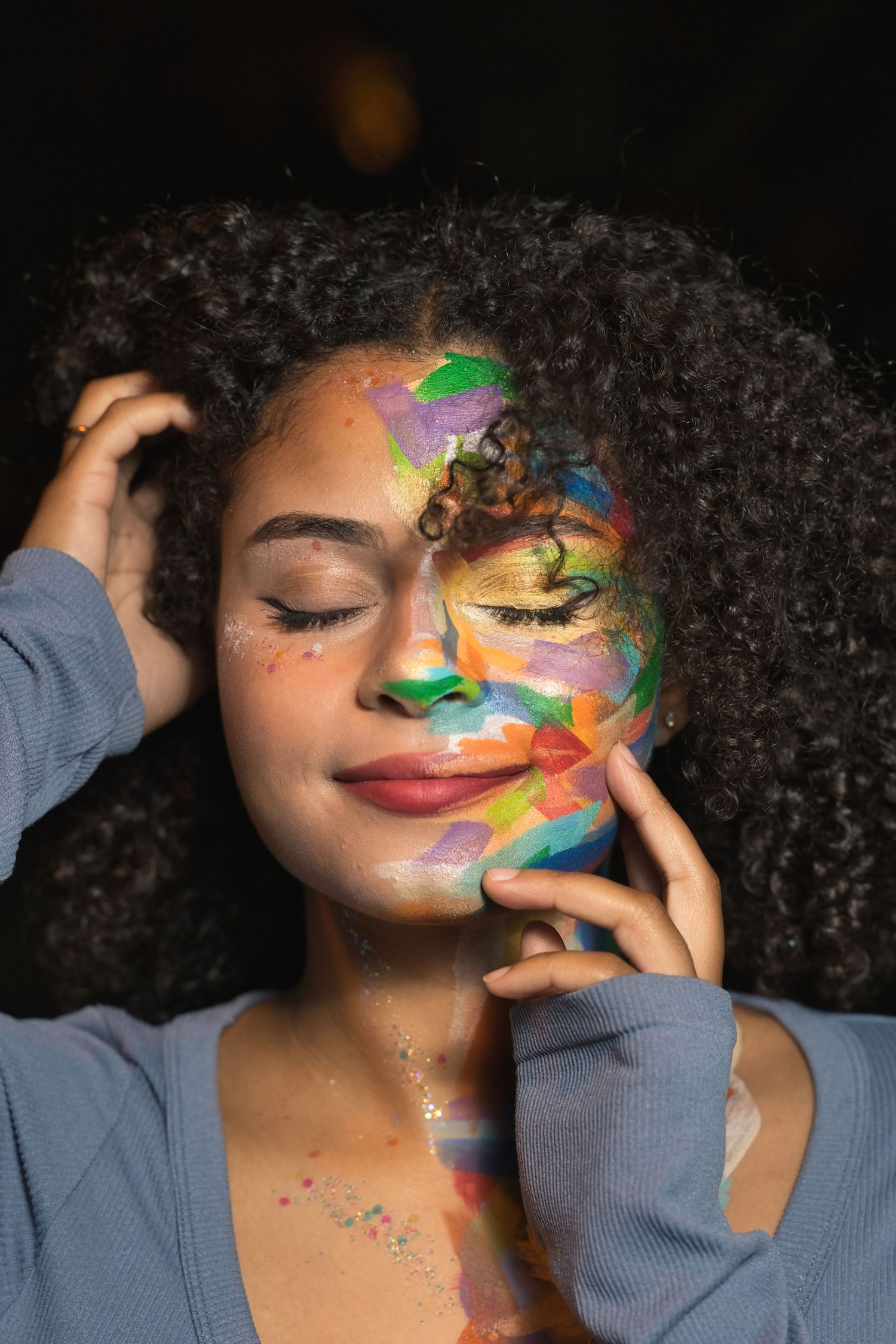 Young woman with curly hair and colorful face paint smiling with eyes closed touching her cheek.