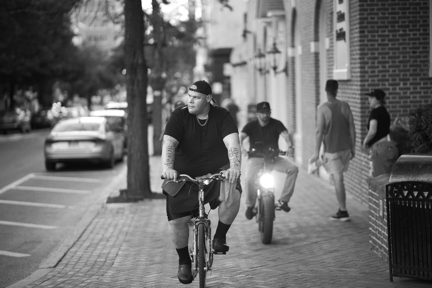 Black and white street photo of people riding bicycles on a brick sidewalk at night.