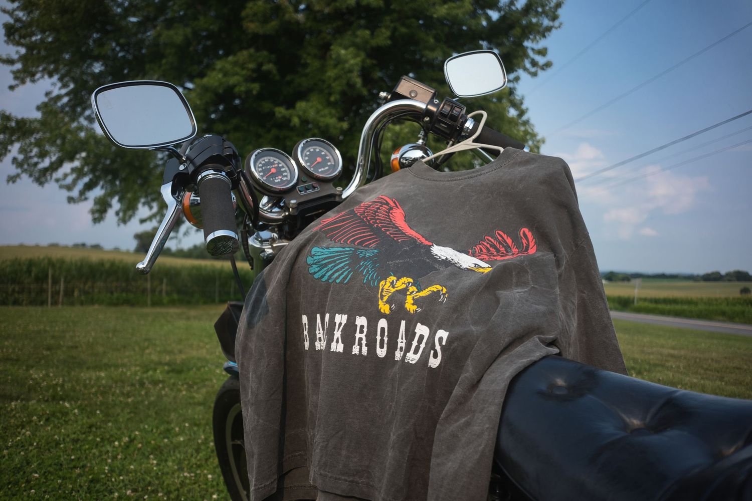 A gray Back Roads graphic t-shirt draped over a classic motorcycle parked on green grass near trees.