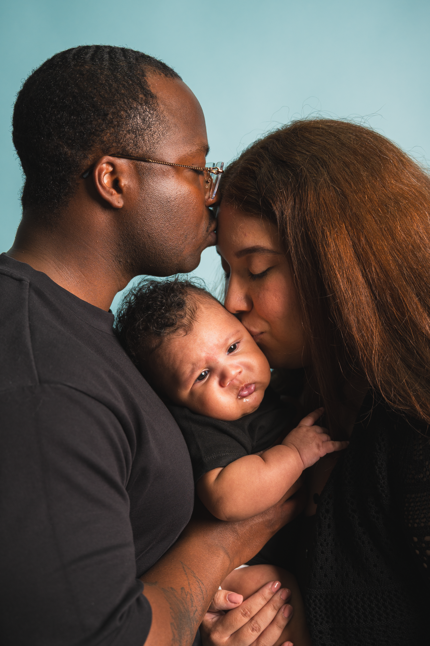 Parents holding their newborn baby in a studio portrait session. Dad kissing mom on the forehead.