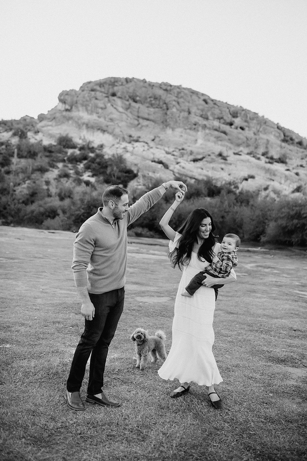 Black and white family photo with couple dancing outdoors, woman holding baby, small dog nearby, rocky landscape background.