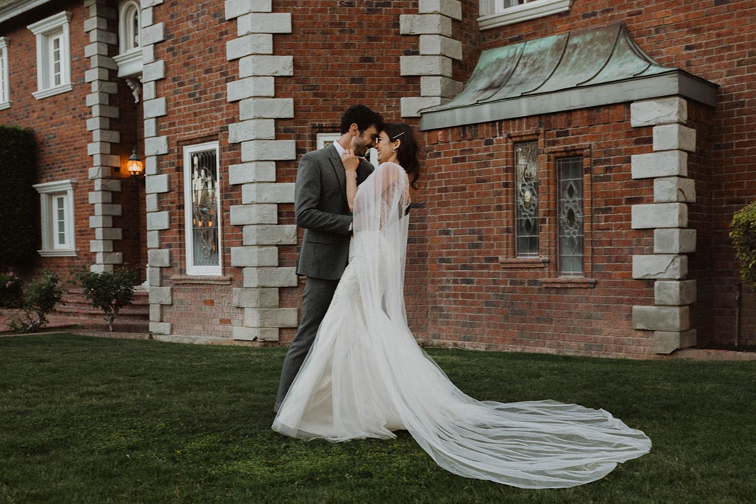 A bride in a flowing white gown with a long cathedral veil stands outside a historic brick building.