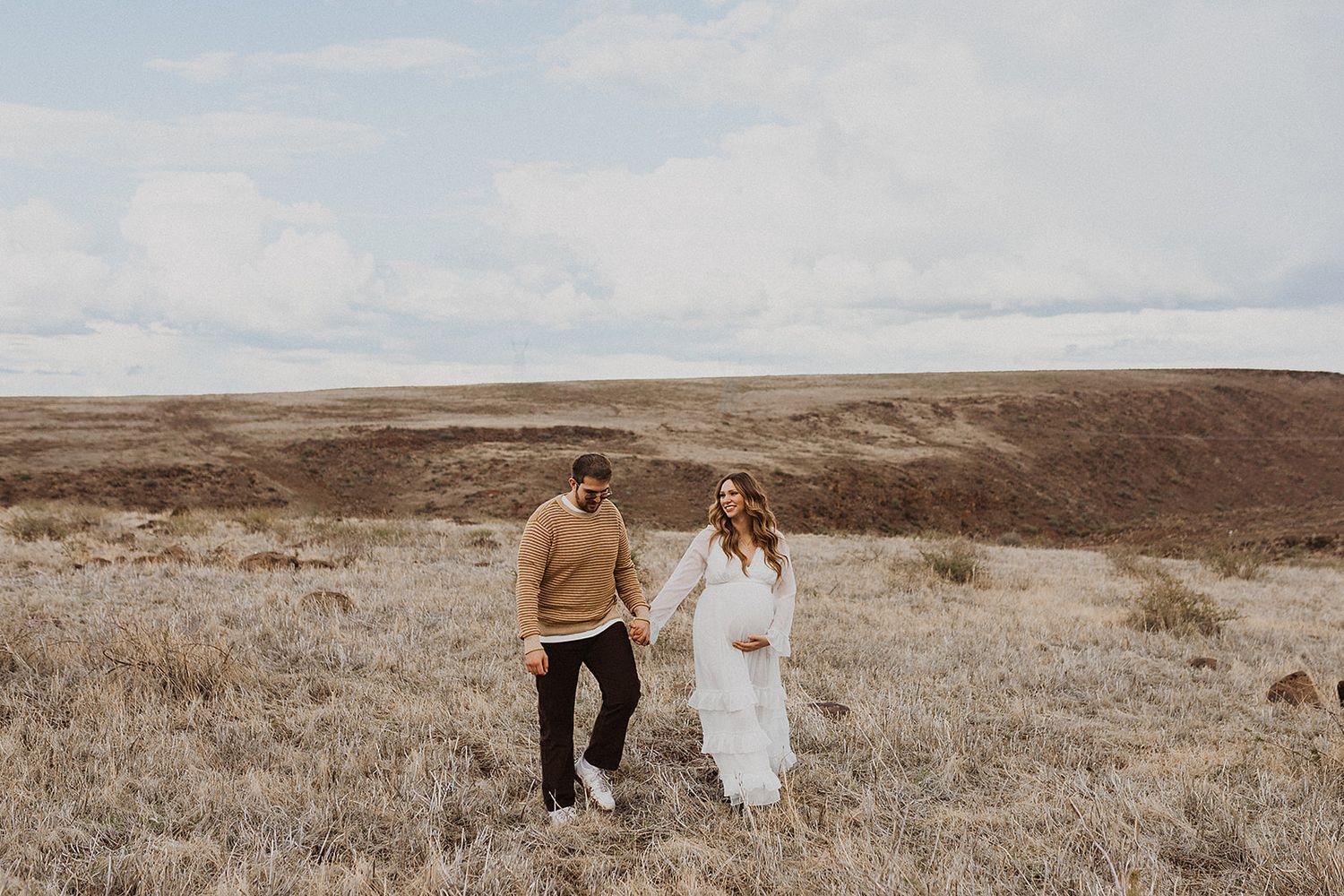 A couple walks hand in hand through a scenic desert landscape during a golden hour photo session.