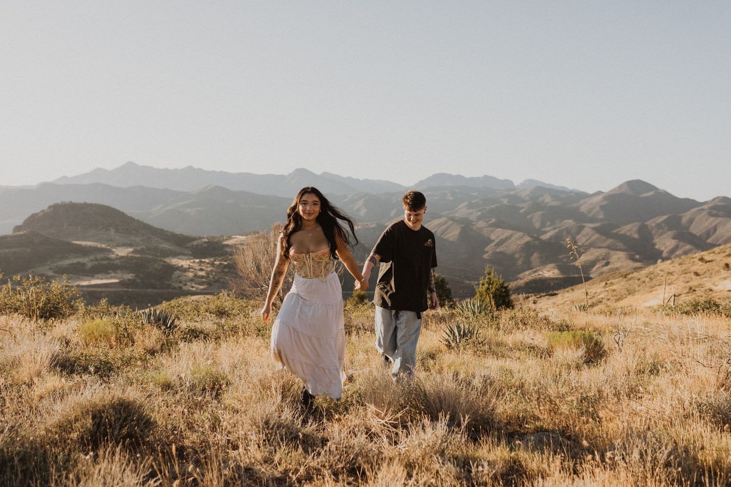 Two people walk hand in hand through golden mountain meadows at sunset with rolling hills in the distance.