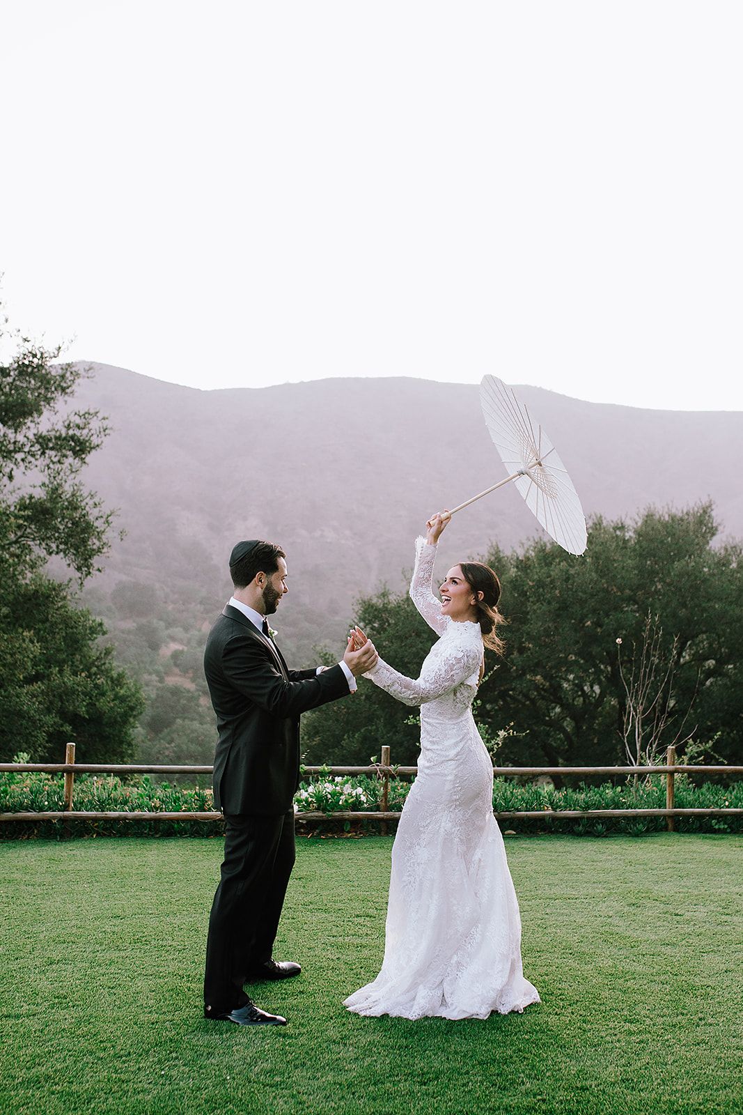 A couple in wedding attire playfully duels with white umbrellas on a grassy lawn with mountains in the background.