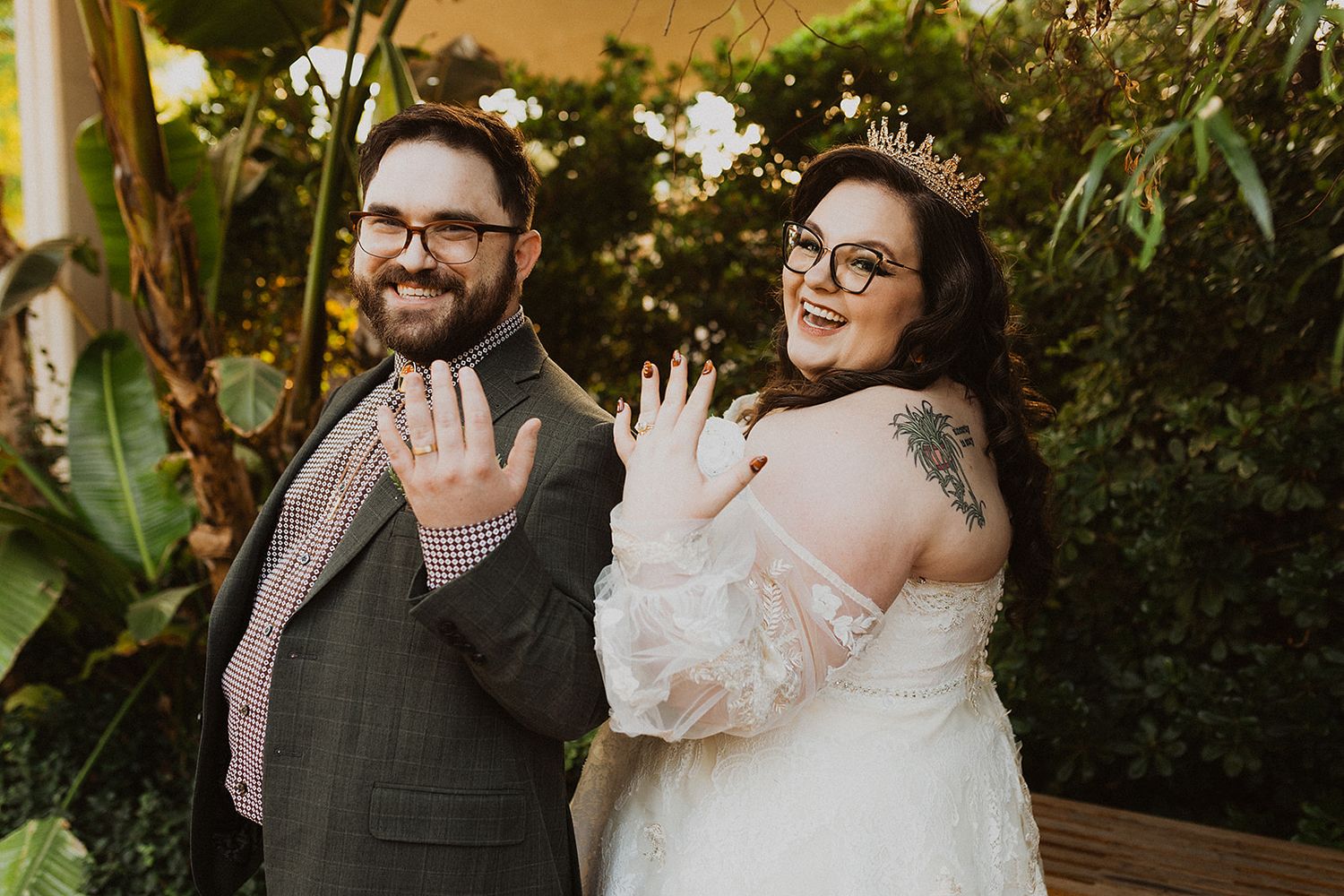 A happy couple in wedding attire showing their rings while standing in a lush garden setting.