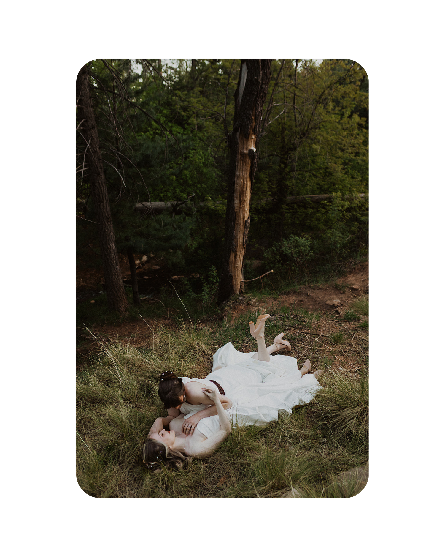 A person in a flowing white dress lies gracefully on the forest floor surrounded by lush greenery and fallen leaves.