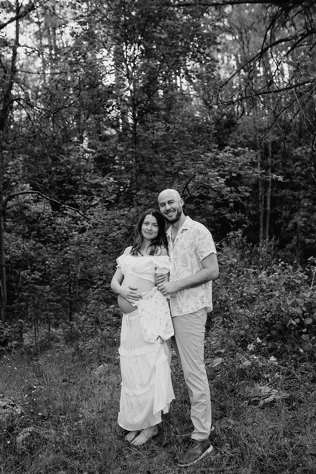 A black and white photograph of a couple embracing in a wooded forest setting.