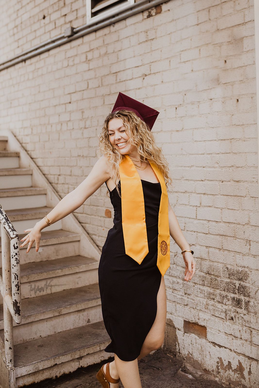 Smiling graduate in black dress, gold stole, and maroon cap poses on outdoor stairs against brick wall.