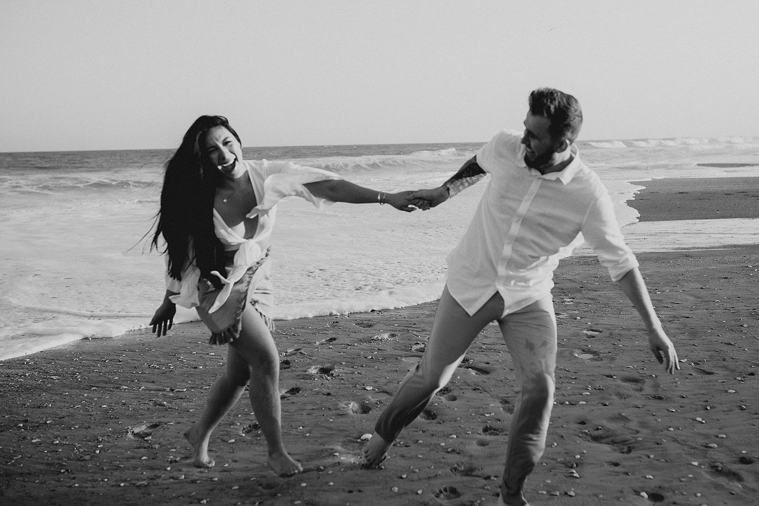 A playful black and white photo of two people dancing and having fun on a beach at sunset.