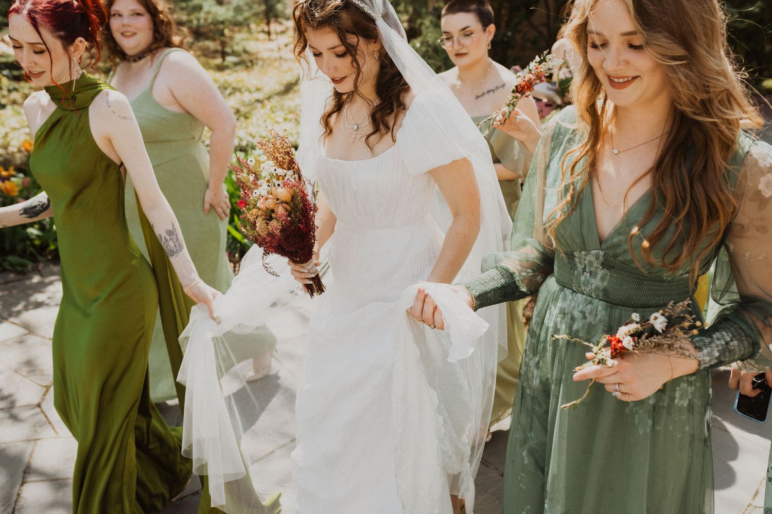 Wedding party in sage green dresses and white gowns celebrate joyfully outdoors during summer celebration.