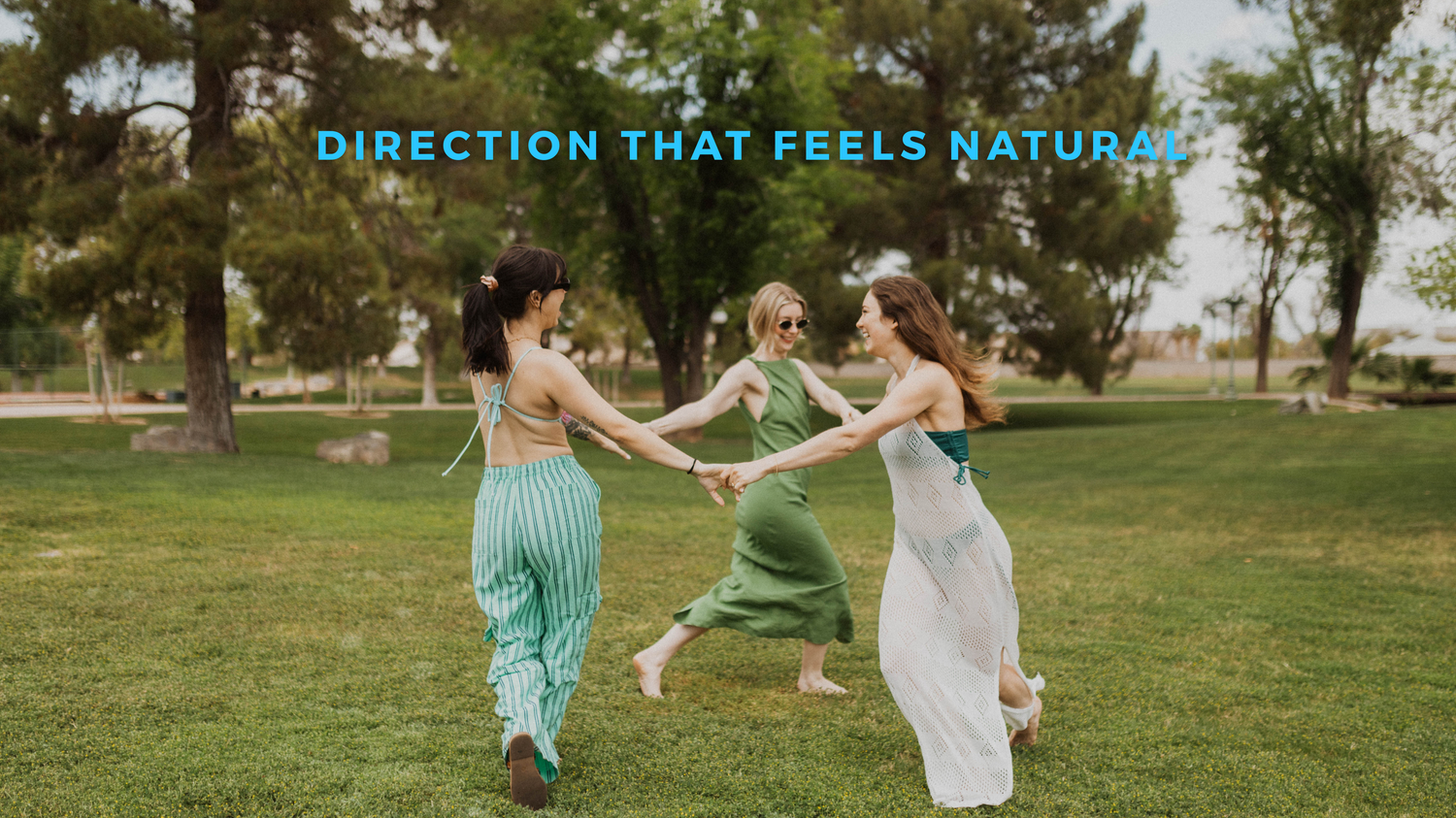 Three friends in flowing dresses dance and hold hands together in a grassy field on a sunny day.