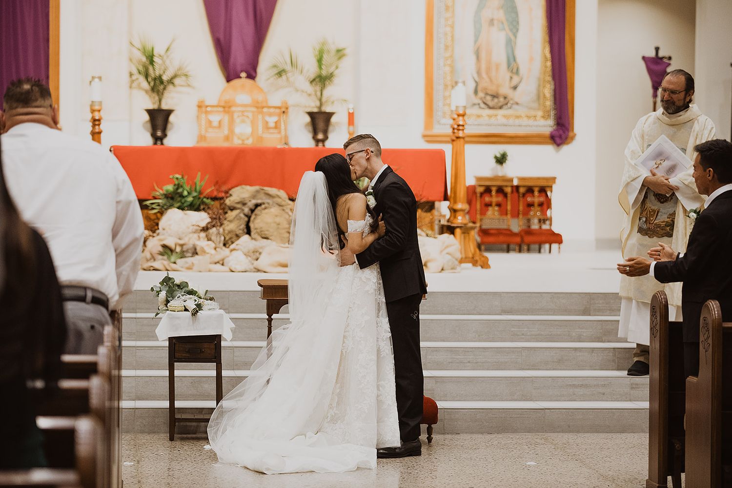 A couple shares their first kiss as newlyweds at the altar of a Catholic church decorated with floral arrangements.
