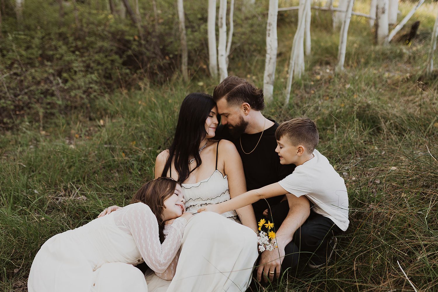 Family of four sitting together in a lush green forest, sharing a tender moment during an outdoor maternity photo session.